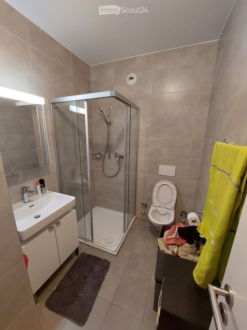 Modern bathroom with a white sink, toilet, glass-enclosed shower, and a yellow towel hanging on the wall. The space features beige tiles and a dark gray bath mat.