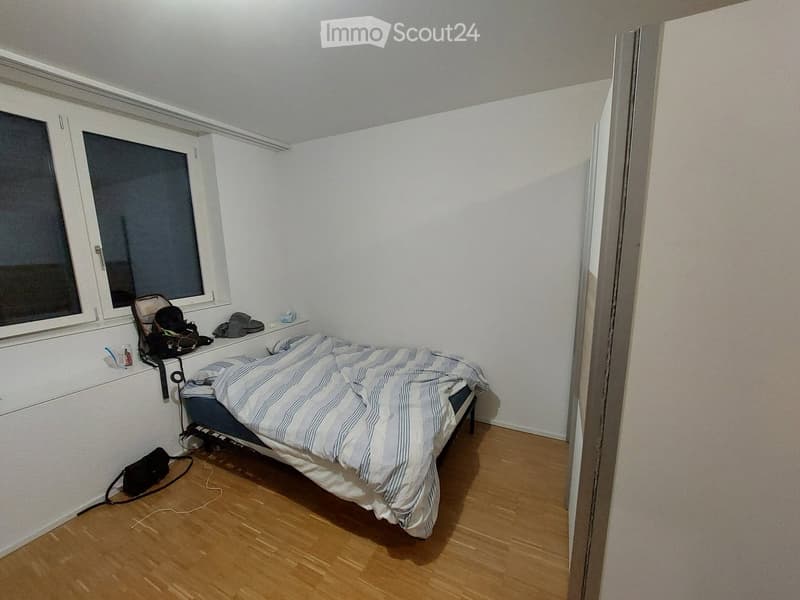A small bedroom with a wooden floor, a single bed with striped bedding, a window with a view, and various personal items on a shelf and the floor.
