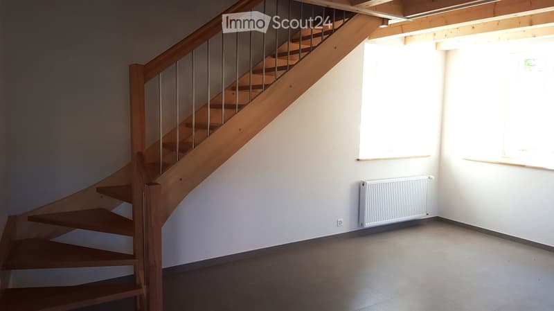 Empty hallway with a wooden staircase, white walls, tiled floor, radiator, and large windows allowing natural light.