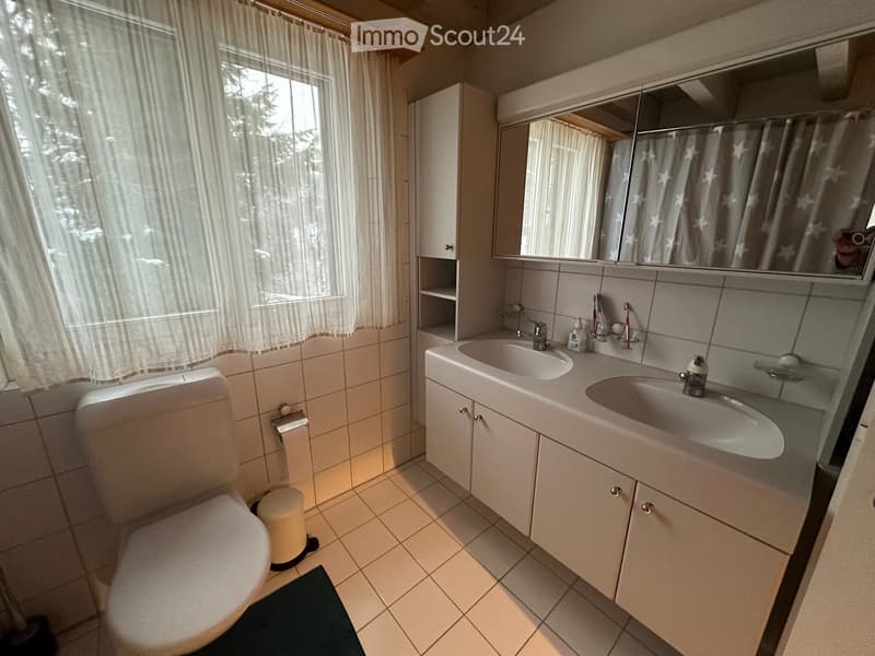 A clean, modern bathroom with a toilet, double sink, mirror, and cabinet. The room features tiled walls and floor, a window with curtains, and a small trash bin near the toilet.