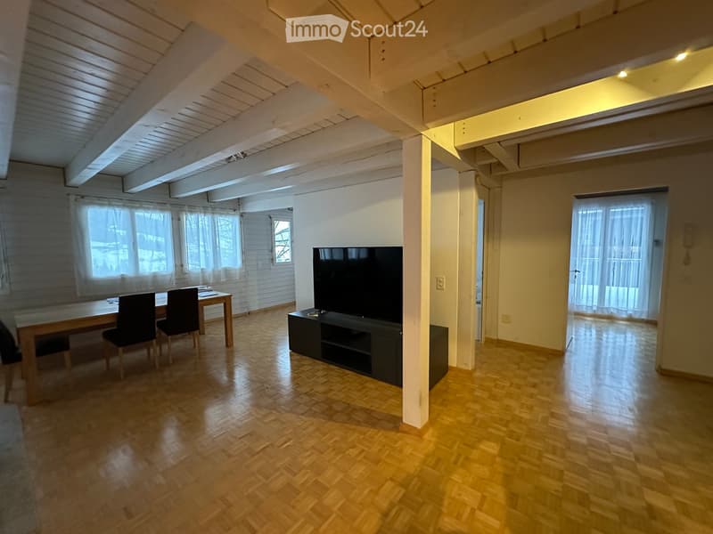 Bright, open living space with wooden flooring, large windows, a TV on a cabinet, a dining table with chairs, and exposed wooden beams on the ceiling.