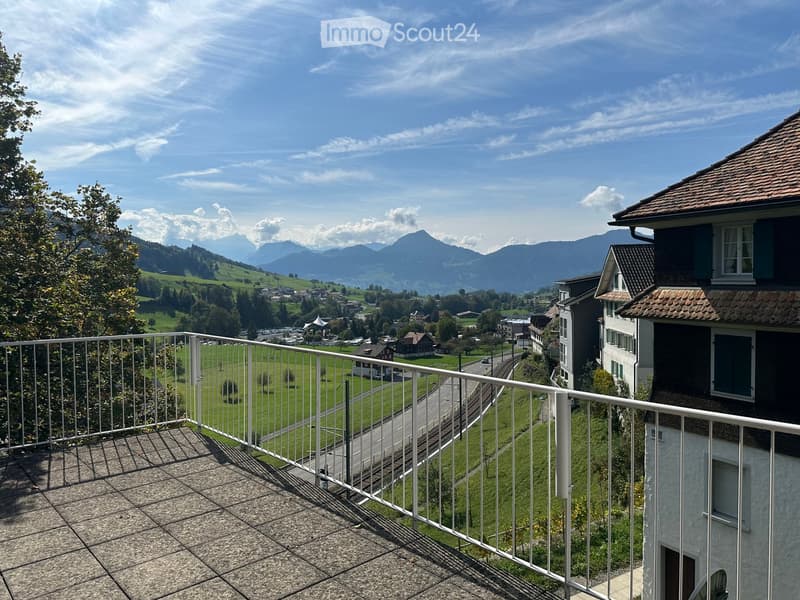 Balcony with metal railing overlooking a scenic valley and distant mountains, with a town and green fields below.