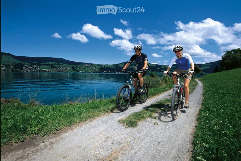 Two cyclists ride along a gravel path beside a lake, surrounded by green hills and a clear blue sky.