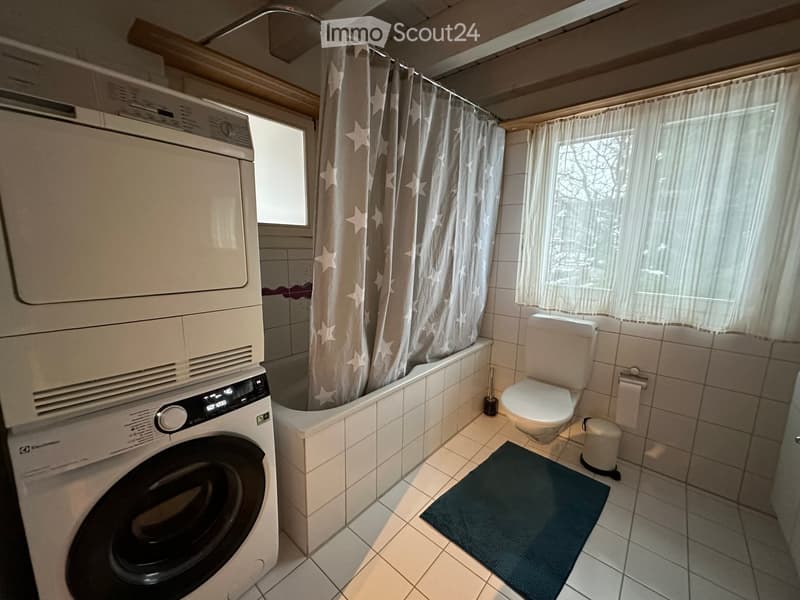 Bathroom with a bathtub, toilet, washing machine, and dryer unit, featuring a shower curtain with star patterns and a small window with sheer curtains.