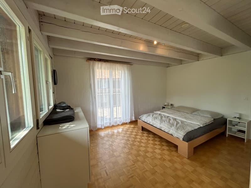 Bright bedroom with wooden flooring, a bed with striped bedding, a nightstand, a window with white curtains, and a mirror on the wall.