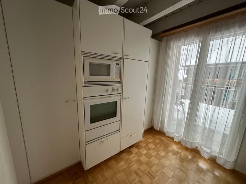 Modern kitchen with light-colored cabinets, a microwave, and an oven, featuring wooden flooring and sheer white curtains on the window.