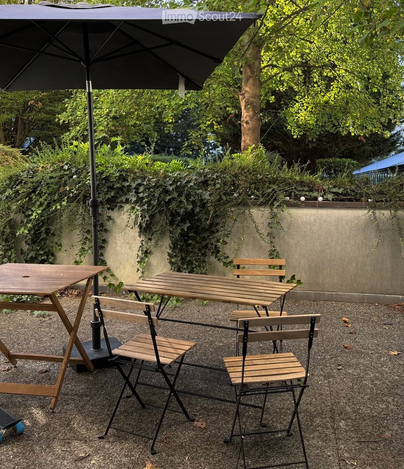 Garden area with gravel ground, two wooden tables, four wooden chairs, and a large black umbrella providing shade.