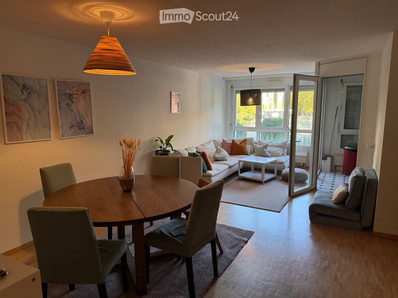 Modern living and dining area with a round wooden dining table, four chairs, a sofa with multiple cushions, a coffee table, a small ottoman, a stool, and a vase with dried flowers on the table. Large windows and a sliding glass door lead to a balcony.
