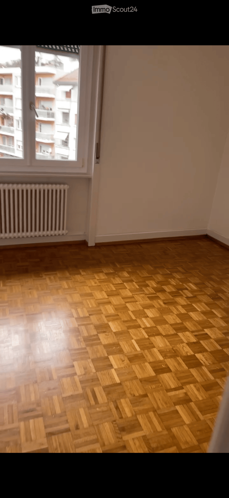 Empty room with wooden herringbone flooring, light-colored walls, and a window with a radiator below it.