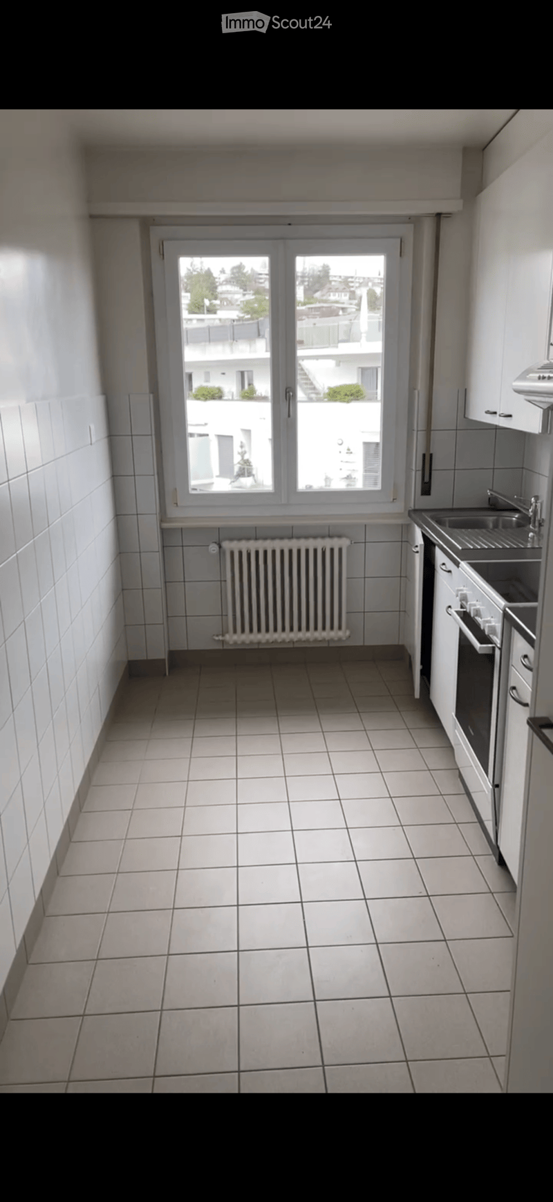 Empty kitchen with tiled floor, white tiled walls, a radiator, a sink, and a window with a view of the building exterior.
