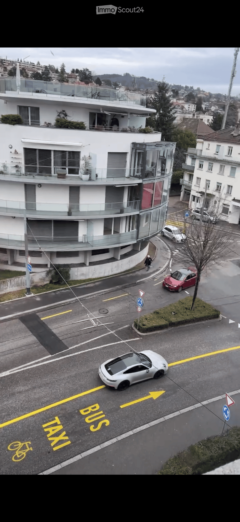 A modern multi-story building with balconies and large windows, surrounded by a street with several parked cars and traffic markings for buses, taxis, and bicycles.