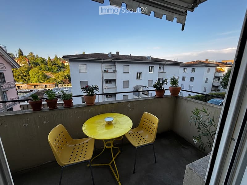 Balcony with yellow table and chairs, potted plants, and a view of surrounding buildings and greenery.