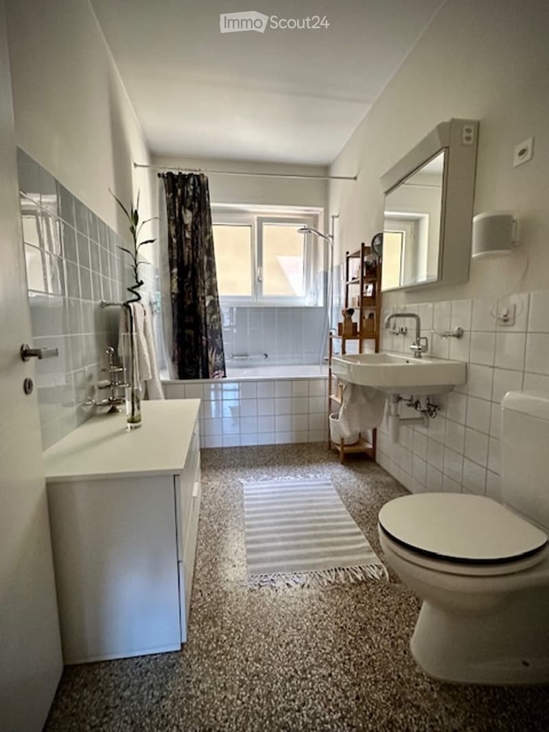 Modern bathroom with white tiles, a sink, toilet, bathtub, and a large mirror above the sink. A small shelf holds towels and a decorative item, and a window with a dark curtain lets in natural light.