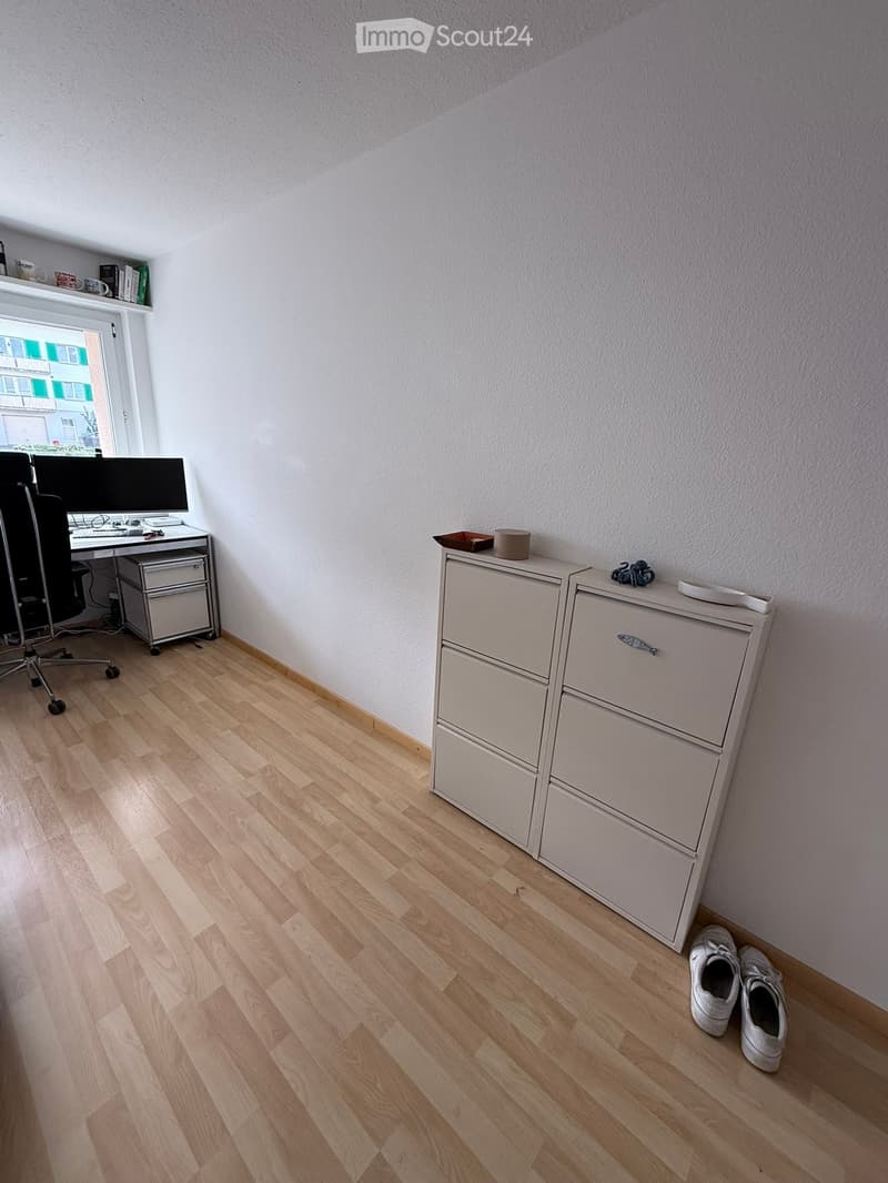 A furnished room with light wooden flooring, a desk with a computer and chair, a filing cabinet, and a pair of shoes on the floor.