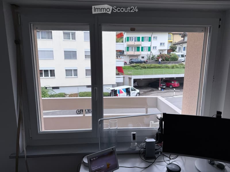 A modern workspace with a laptop, screen, headphones, and laptop stand on a white desk, overlooking a balcony with a view of a residential area and parked cars.