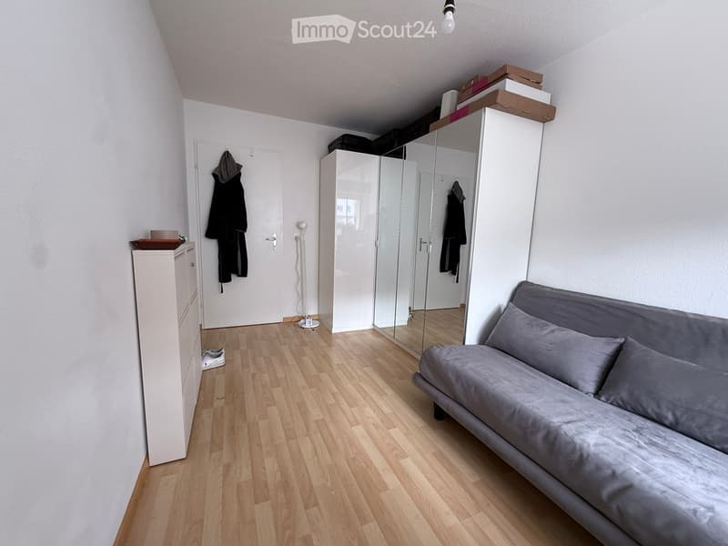 A modern, furnished room with a grey sofa, a large mirrored wardrobe, a white dresser, a coat rack, and a lamp. The room features wooden flooring and white walls.