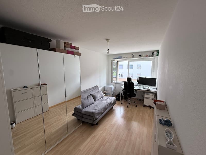 A small, well-lit room with a sofa bed, desk, chair, and computer. The room features wooden flooring, a large window, and a mirror on the wall.