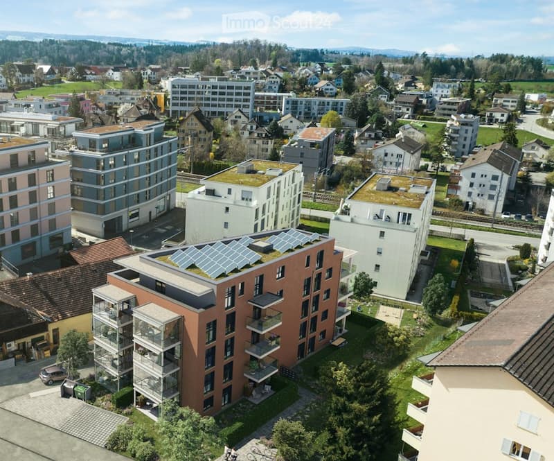 Aerial view of a modern residential complex with multiple buildings, green roofs, and solar panels. The area includes a mix of contemporary and traditional houses, surrounded by trees and open spaces.