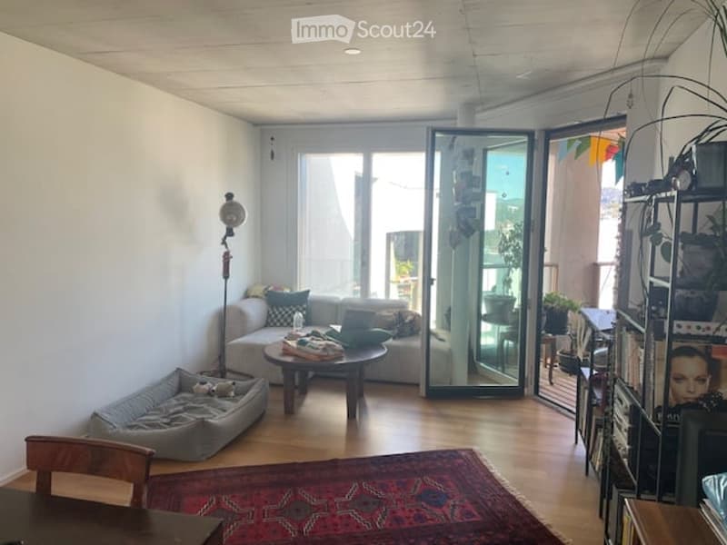 Modern living room with a grey sofa, coffee table, dog bed, floor lamp, and large sliding glass doors leading to a balcony. The room features wooden flooring and a colorful rug.