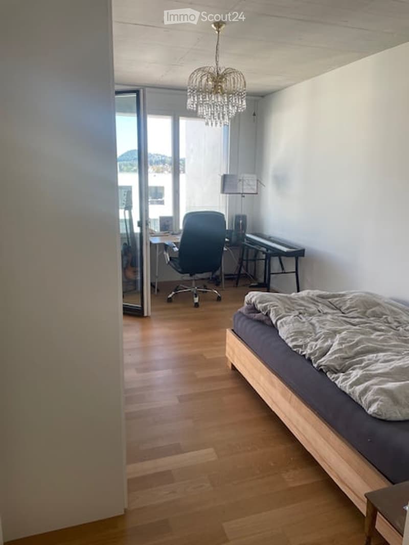 A modern, well-lit room with a bed, desk, and chair. The room features a chandelier, a view through large windows, and a wooden floor.