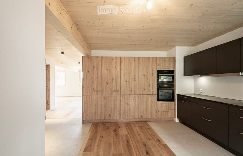 Modern kitchen with wooden cabinets, dark lower cabinets, and a built-in oven. The space features exposed wooden beams on the ceiling and a light wooden floor.