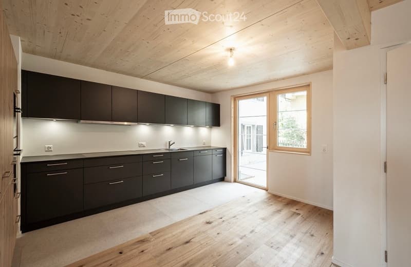 Modern kitchen with dark grey cabinets, wooden flooring, and a large window with a sliding door leading outside.
