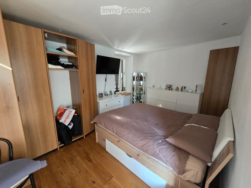Modern bedroom with a wooden bed, wardrobe, dresser, mirror, and television mounted on the wall. The room features wooden flooring and a cozy bed with brown bedding.