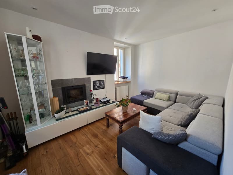 Modern living room with a grey sectional sofa, wooden coffee table, fireplace, flat-screen TV, and glass cabinet. The room features wooden flooring and white walls.