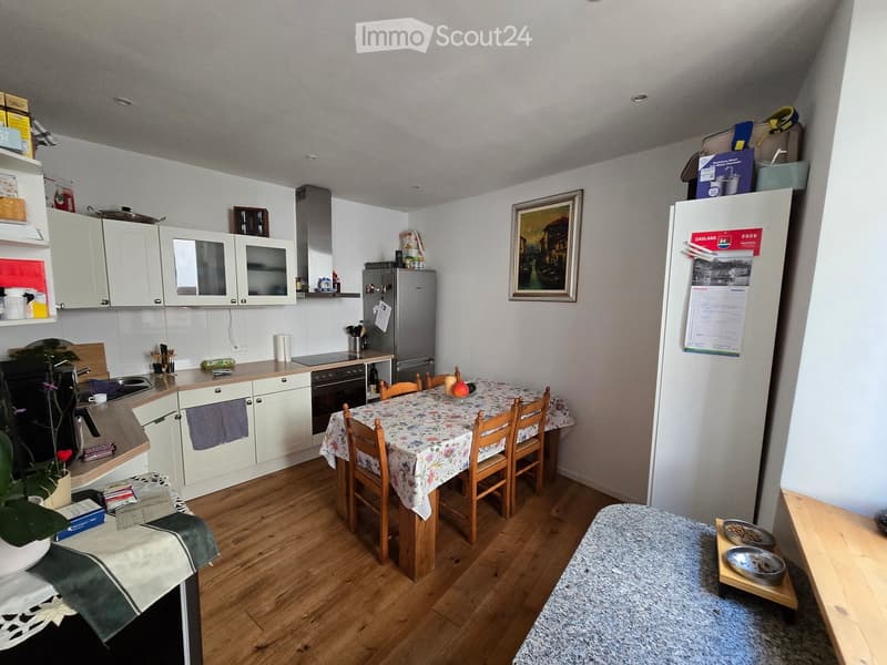 A modern kitchen with white cabinets, a wooden dining table with chairs, a refrigerator, oven, microwave, and a sink. There is a plant on the counter and a picture frame on the wall.