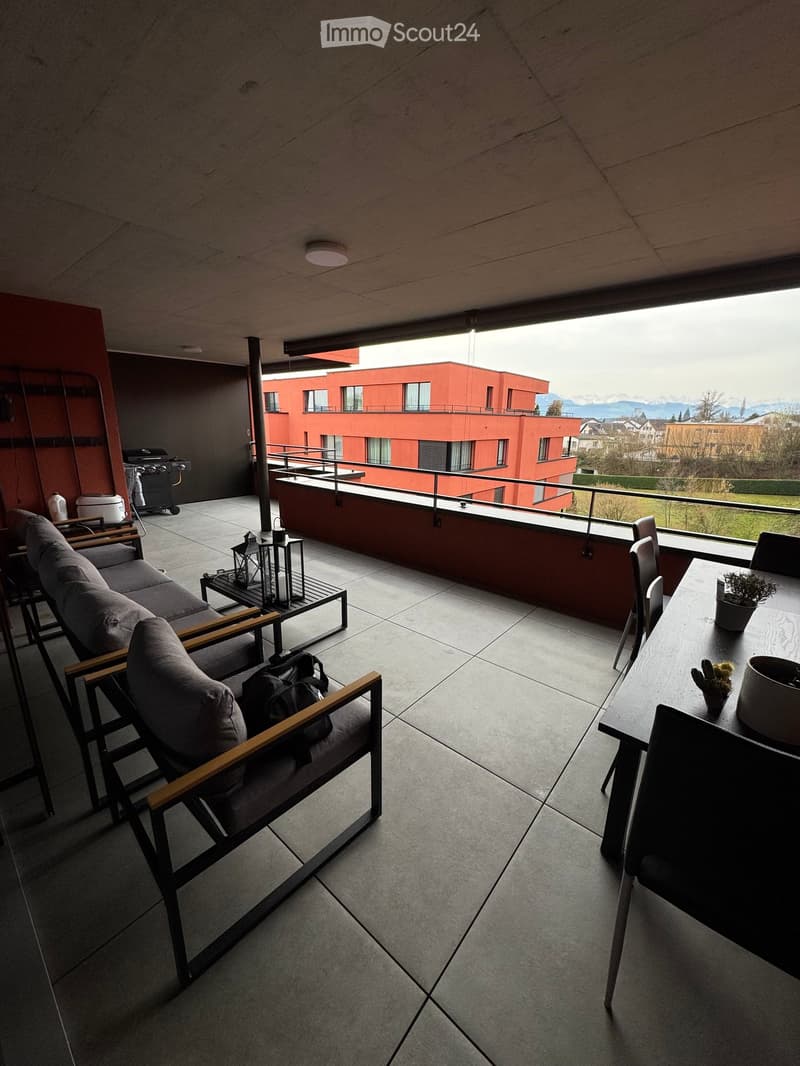 Modern balcony with grey sofa, wooden-framed chairs, small table, lanterns, and a view of a red building and mountains.