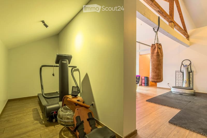 Well-lit fitness room with wooden flooring, featuring a treadmill, elliptical machine, punching bag, and fitness mat.