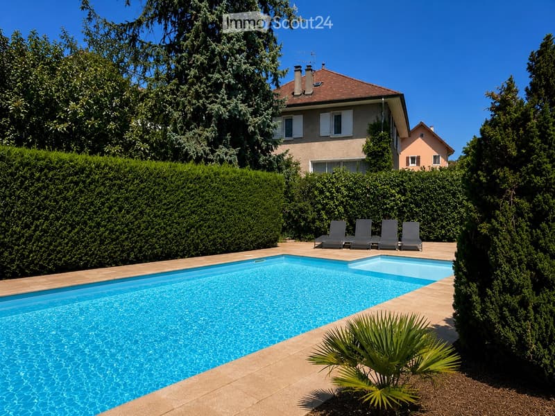 A rectangular swimming pool with clear blue water, surrounded by a tiled deck, lounge chairs, and lush greenery including tall trees and shrubs.