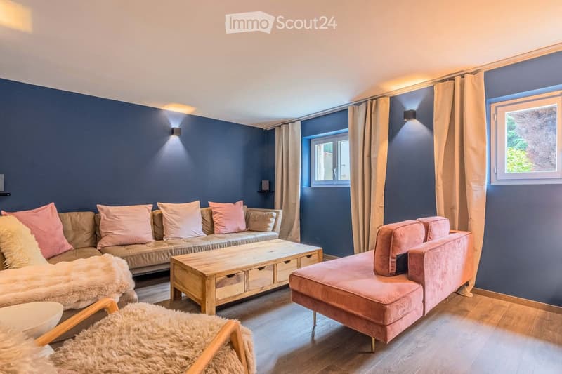 Modern living room with dark blue walls, large windows, and natural light. Features a beige sofa with pink cushions, a wooden coffee table, a pink ottoman, and a fur-covered chair.