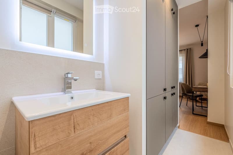 Modern bathroom with a white sink, wooden cabinet, and a mirror above it. The adjacent room features a wooden floor, a chair, and a small table.