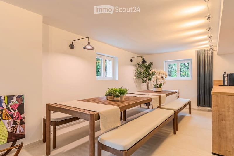 Bright dining area with wooden tables, white benches, and potted plants. Features modern lighting and large windows with a view of greenery.