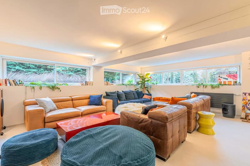 Modern living room with large windows, leather sofas, colorful cushions, a red coffee table, and a blue ottoman.
