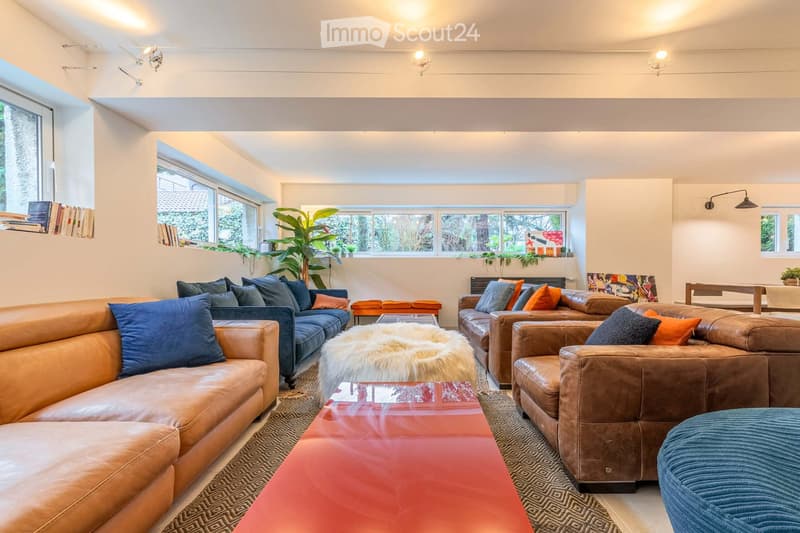 Modern living room with large windows, leather sofas, colorful cushions, a fur throw, a red coffee table, and a patterned rug.