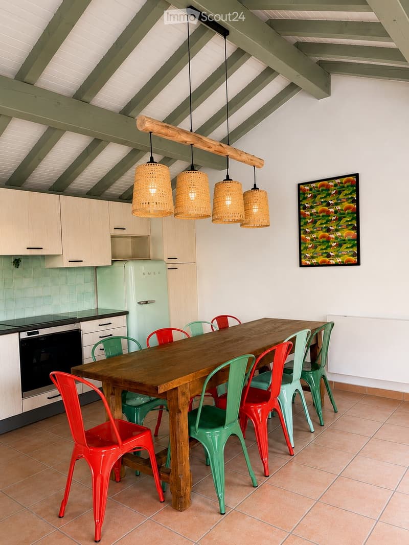 Bright kitchen with a wooden dining table surrounded by colorful chairs, modern cabinets, a vintage refrigerator, and pendant lights hanging from a slanted ceiling.