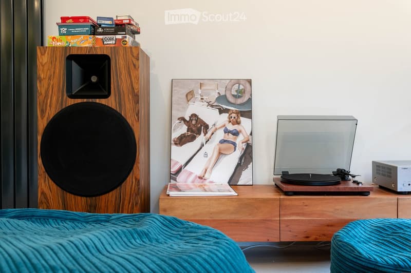 A modern bedroom with a wooden speaker cabinet, a turntable, a large picture frame, and a bed with a blue blanket.
