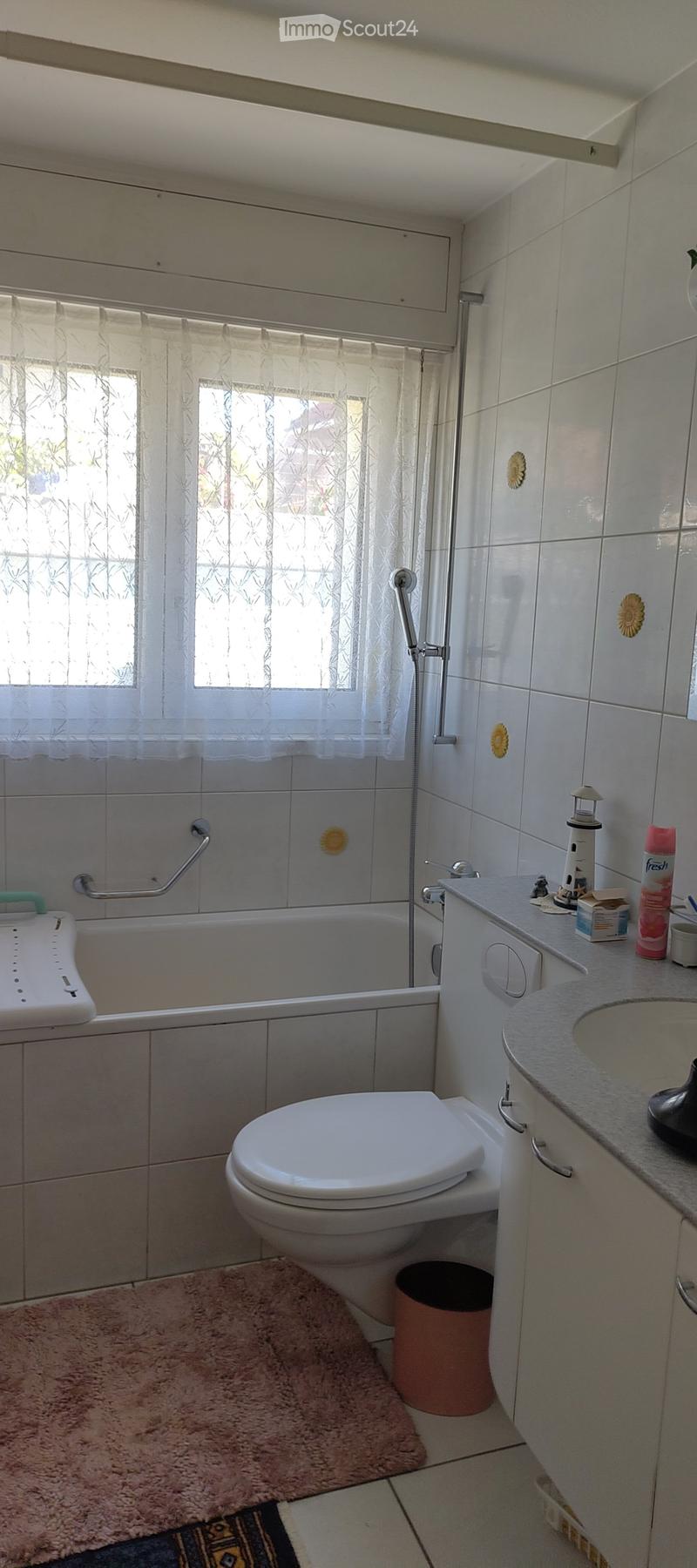 Bathroom with a bathtub, toilet, sink, and cabinet. Features a window with a curtain, a handrail, and a trash can.