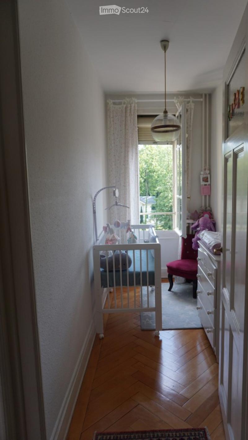 A small, well-lit room with wooden flooring, a crib, a red chair, a dresser, and toys visible. The room has a view of a balcony through an open door.