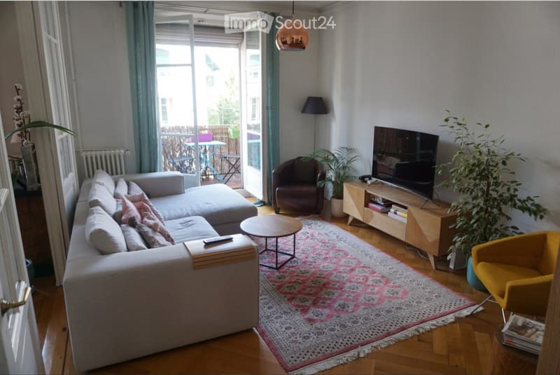 Modern living room with a grey sectional sofa, wooden coffee table, armchairs, TV, and large windows leading to a balcony.