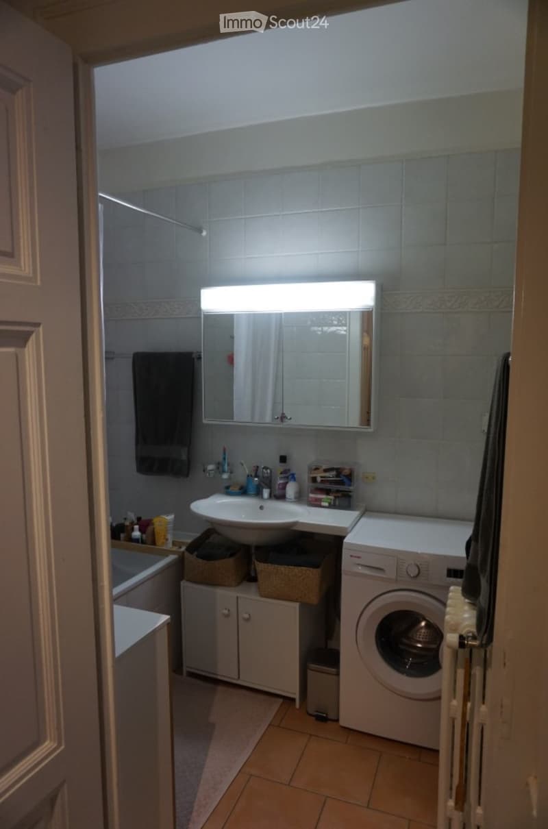Bathroom with a sink, mirror, washing machine, and a small cabinet. The room features tiled walls, a wooden floor, and a towel hanging on the wall.