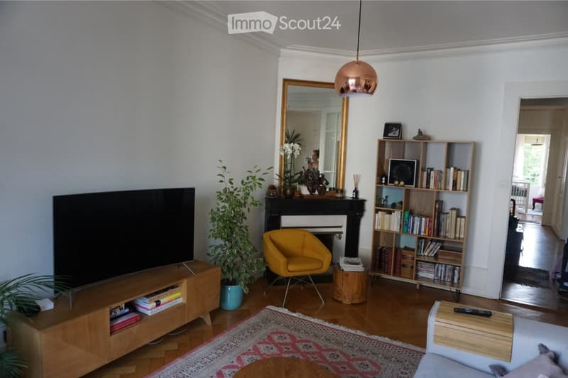 Modern living room with a large television, a yellow armchair, a wooden coffee table, a bookshelf filled with books, a decorative mirror, and a fireplace.