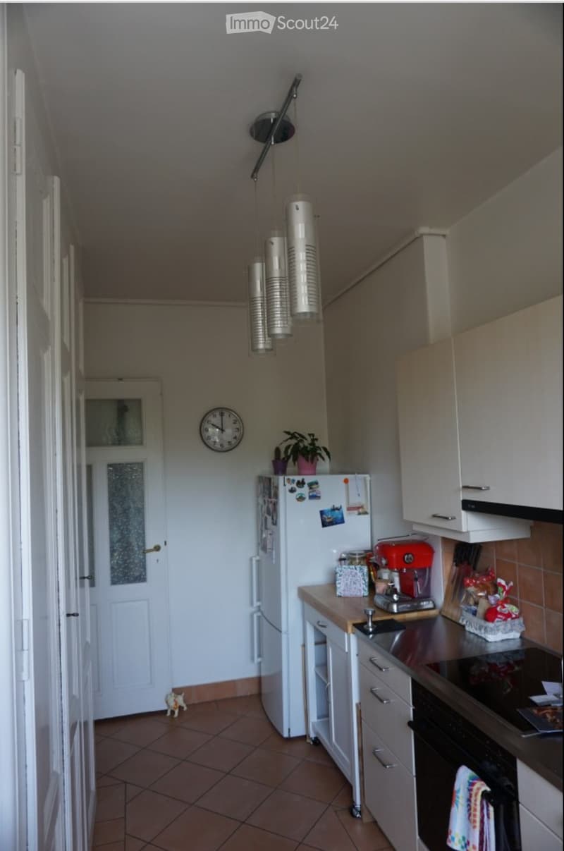 A modern kitchen with white cabinets, a refrigerator, a stove, and a sink. The space features a tiled floor and a hanging light fixture.