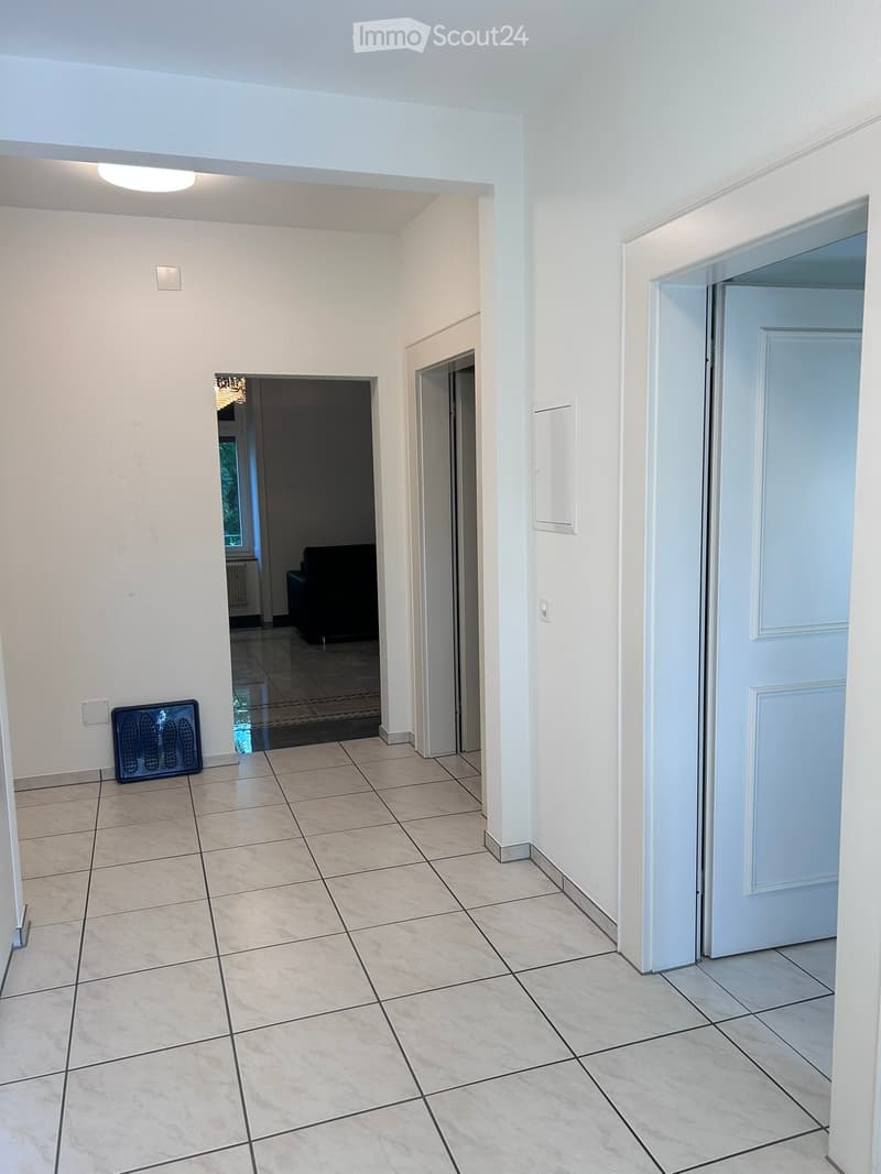 Empty hallway with tiled floor, white walls, and several doors leading to other rooms. A radiator is visible on the left wall, and a light fixture is mounted on the ceiling.