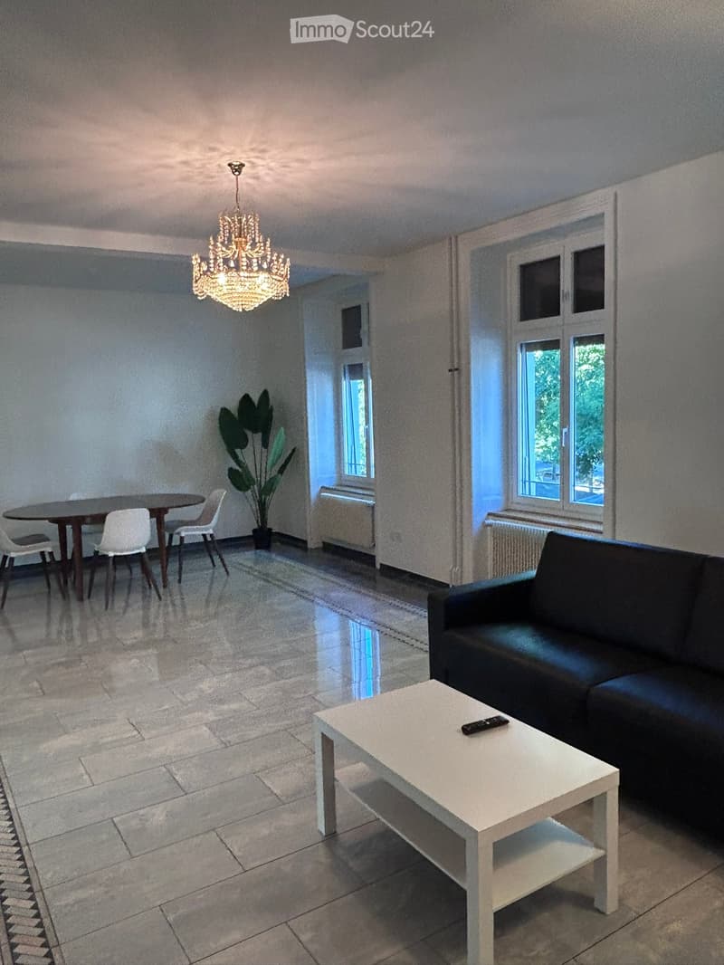 Living room with a black leather sofa, white coffee table, dining table with chairs, chandelier, and a potted plant.