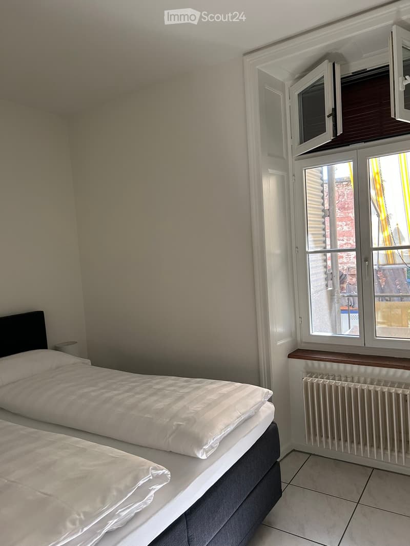 Cozy bedroom with two single beds, white bedding, tiled floor, radiator, and large window with view.