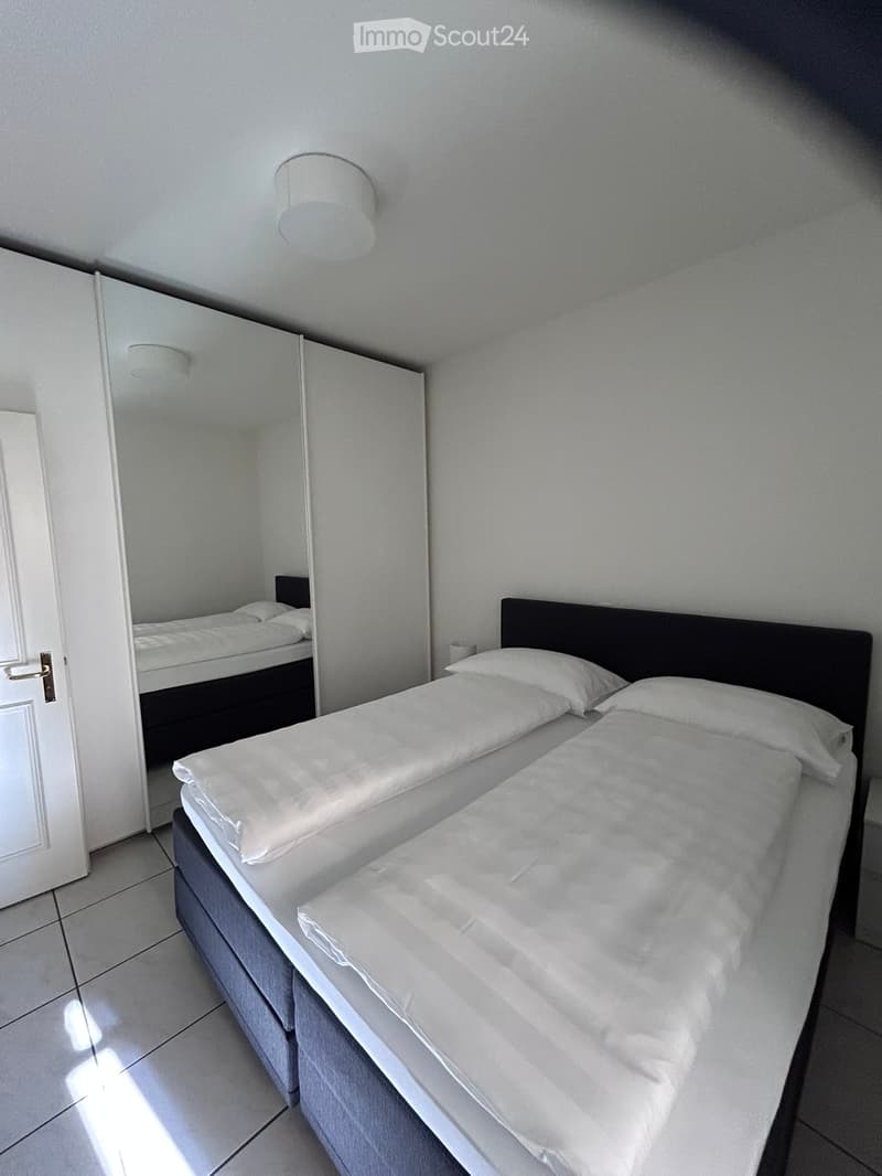 A modern, minimalist bedroom with a double bed, white bedding, a wardrobe, and a large mirror reflecting another bed.