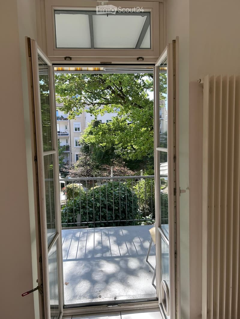 Open sliding glass door leading to a small balcony with wooden decking, metal railing, and greenery visible outside.